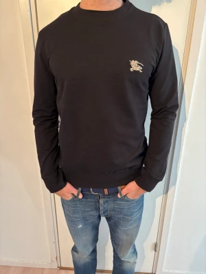 Burberry sweatshirt   - Stilren svart sweatshirt från Burberry med rund halsringning och diskret Burberry-logga broderad på bröstet. Tröjan har ribbade muddar vid ärmslut och nederkant. Perfekt för en clean och klassisk look.
