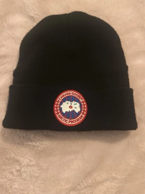 Svart mössa från Canada Goose - Säljer en svart ribbstickad mössa från Canada Goose med uppvikt kant och klassisk rund logotyp-patch framtill i rött, vitt och blått. Perfekt för kalla dagar och enkel att matcha med olika outfits.