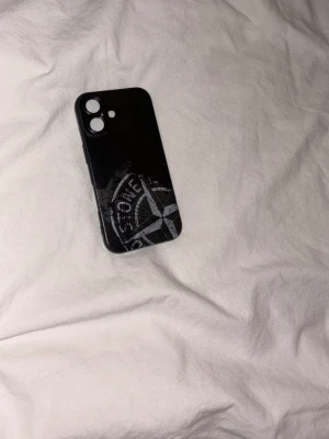 Svart mobilskal med Stone Island-tryck - Snyggt svart mobilskal med Stone Island-kompass-tryck i grått och vitt. Skalet är gjort i tålig plast och har öppningar för kamera och knappar. Passar perfekt för dig som vill ha en stilren och trendig look på mobilen. iPhone 16