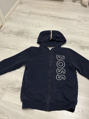 Mörkblå hoodie från BOSS - Snygg mörkblå hoodie från BOSS med dragkedja framtill och stor vit BOSS-logga längs bröstet. Huva med dekorativ rand i beige och vitt. Tillverkad i mjukt bomullsmaterial, perfekt för en avslappnad och trendig look.