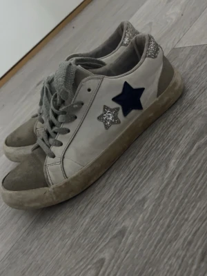 Vita sneakers med glitterstjärnor - Snygga vita sneakers med grå detaljer, glittriga silverstjärnor och en mörkblå stjärna på sidan. Skorna har grå snören och glitter på hälen . Bli dom sålda så gör jag de så rena som möjligt! Pris går att diskuteras!