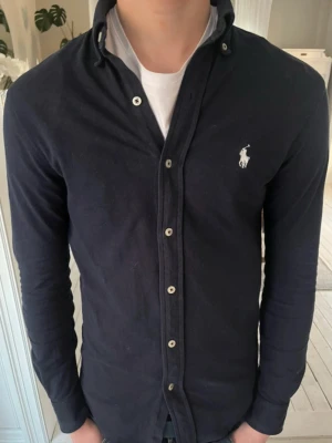 Mörkblå skjorta från Ralph Lauren - Klassisk mörkblå skjorta från Ralph Lauren i featherweight mesh. Perfekt till både jeans och chinos för en clean look. Storlek XS. Modell är 174