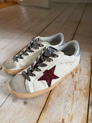 Golden Goose sneakers med röd stjärna - Snygga sneakers från Golden Goose i vitt läder med silverfärgad häl, grå mockadetaljer på tån och leopardmönstrade skosnören. På sidan sitter en röd glittrig stjärna. Yttersulan är beige.