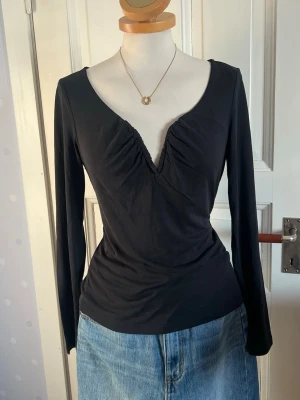Svart långärmad topp med v-ringning - Svart långärmad topp med djup v-ringning och rynkade detaljer vid bysten. Figurnära passform och mjukt, stretchigt material som ger en snygg siluett. Perfekt att styla med jeans eller kjol.