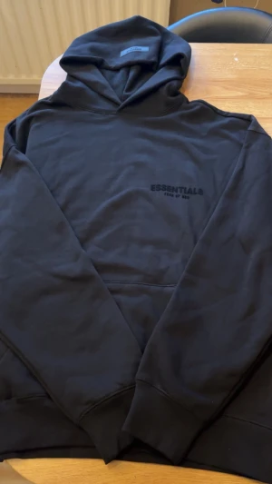 Svart Essentials Fear of God hoodie - Säljer en svart hoodie från Essentials Fear of God med diskret logga på bröstet. Hoodien har klassisk passform, stor ficka framtill och ribbade muddar. Tillverkad i mjukt bomullsmaterial som är perfekt för en avslappnad streetwear-look.