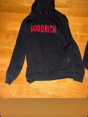 Svart HOODRICH hoodie med röd text - Svart hoodie från HOODRICH med stor röd logga på bröstet. Klassisk känguruficka framtill och justerbar huva med snören. Perfekt för en chill och streetwear-inspirerad look. Tillverkad i mjukt material för extra komfort.