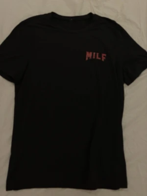 Svart t-shirt med flamingotryck - Svart t-shirt med rött tryck fram och stort motiv på ryggen med texten 'MILF - Man I Love Flamingos' och en flamingo i solnedgång. Klassisk passform och rund hals. Perfekt för dig som gillar statement-plagg och coola prints. Det står S men är mer en M