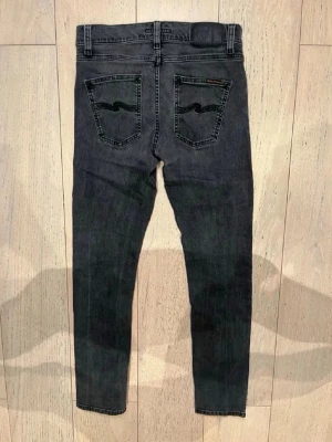 Svarta slim jeans från Nudie Jeans - Säljer ett par svarta slim jeans från Nudie Jeans med klassisk femficksmodell och diskreta sömmar på bakfickorna. Jeansen har en smal passform och är gjorda i stretchigt denimtyg för extra komfort. Perfekta för en avslappnad och trendig look.