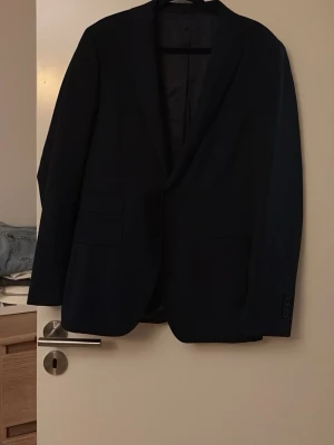 Svart kavaj från These Glory Days - Stilren svart kavaj från These Glory Days med klassisk passform och två framfickor. Materialet är en mix av ull och polyester vilket ger en skön känsla och bra hållbarhet. Perfekt för dig som vill ha en tidlös och snygg look.