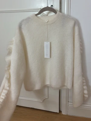 Vit fluffig stickad tröja med volangärm - Super populär vit stickad tröja med volangdetaljer längs ärmarna. Tröjan har en  rund halsringning och är gjord i ett mjukt, fluffigt material som känns lyxigt. Perfekt för dig som vill ha något extra till din outfit.