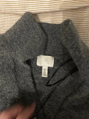 Grå stickad tröja från H&M - Mysig grå stickad tröja från H&M med hög krage och långa ärmar. Tröjan har ribbstickade muddar vid ärmslut och nederkant, vilket ger en snygg och avslappnad look. Perfekt för kyliga dagar och enkel att matcha med jeans eller kjol. Den är nopprig