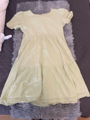 Ljusgrön kort klänning med spetskant - Söt ljusgrön klänning med korta puffärmar och rund halsringning. Klänningen har volangdetaljer och spetskant längst ner, vilket ger en romantisk känsla. Tillverkad i ett lätt tyg, perfekt för varmare dagar.