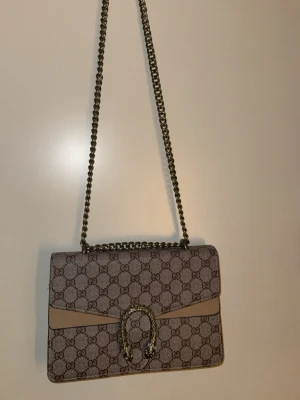 Mönstrad axelväska beige kedja - Supersnygg mönstrad axelväska i beige med klassiskt GG-mönster och silverfärgad kedjerem. Väskan har ett dekorativt spänne , en orm framtill och är tillverkad i canvas med läderdetaljer. Perfekt för dig som vill ha en ikonisk , lyxig, trendig väska. Den har flera fack, se bilder på innehållet. Supersnyggt. Populär o kommer läggas ut på fler sidor,så skynda om ni är sugna på denna supersnygga classy väskan 👜👛