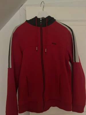 Röd hoodie med dragkedja från BOSS - Snygg röd hoodie från BOSS med svart huva och dragkedja framtill. Har vita och svarta ränder längs ärmarna och diskret BOSS-logga på bröstet. Utrustad med dragsko i huvan och två fickor framtill. Perfekt för en sportig och avslappnad stil.