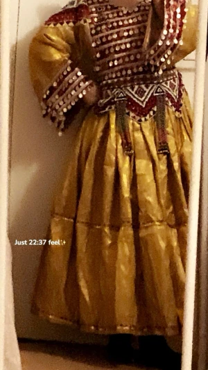 Traditionell gul klänning med speglar - Unik traditionell klänning i gult tyg med vida ärmar och dekorativa speglar och pärlor på bröst och ärmslut. Överdelen har röda och vita detaljer samt ett broderat midjeband med geometriska mönster och hängande dekorationer. Perfekt för dig som vill sticka ut. Har sjal och byxa. 