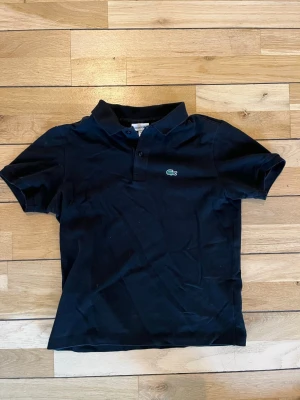 Svart pikétröja från Lacoste - Klassisk svart pikétröja från Lacoste med den ikoniska gröna krokodilloggan på bröstet. Tröjan har korta ärmar, krage och knappar vid halsen. Perfekt till en avslappnad men ändå stilren look.