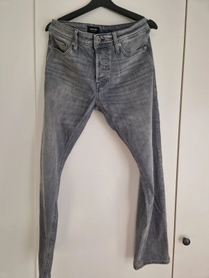 Grå raka jeans från Jack & Jones - Säljer ett par grå jeans från Jack & Jones med rak passform och klassisk femficksdesign. Jeansen har en snygg tvättad look och är gjorda i denim med metallknappar och dragkedja. Perfekta för en avslappnad stil.