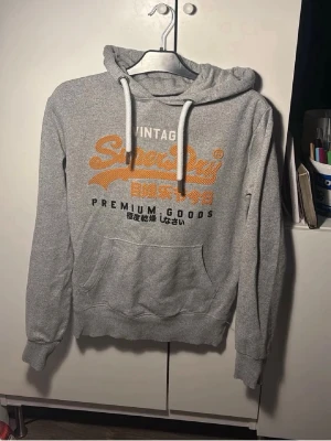 Grå Superdry hoodie med tryck - Säljer en grå hoodie från Superdry med orange och svart tryck på bröstet där det står 'Vintage Superdry Premium Goods'. Hoodien har huva med vita snören och en stor magficka. Perfekt för en avslappnad och cool stil.