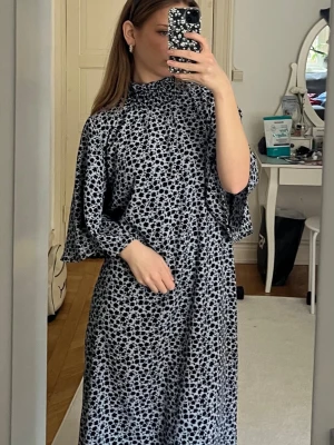 Blommig långklänning med vida ärmar - Säljer en svartblå långklänning med blommigt mönster och vida, trekvartsärmar. Klänningen har hög hals och en loose passform som ger en avslappnad och cool look. Perfekt för dig som gillar att sticka ut med mönster och komfort.