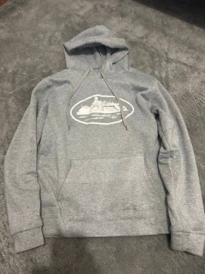 Corteiz hoodie  - Säljer en grå hoodie med vit tryckt grafik på bröstet och klassisk magficka. Hoodien har dragsko i huvan och ribbade muddar vid ärmslut och nederkant. Perfekt för en avslappnad och chill stil.