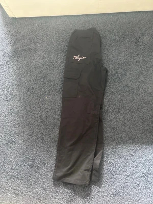  True Religion - Säljer ett par svarta cargobyxor från True Religion med broderad logga på benet och bakfickor med den klassiska vita sömmen. Byxorna har flera fickor och är tillverkade i ett slitstarkt bomullsmaterial. Perfekta för en streetwear-look.