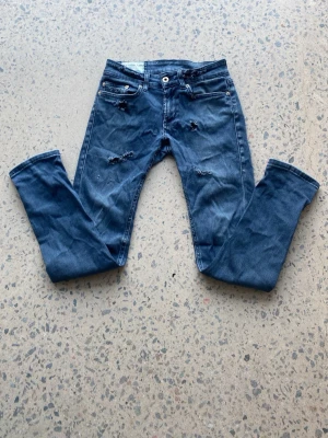 Blå slitna jeans från Dondup - Säljer ett par blå  jeans med slitningar och detaljerade rivna partier framtill. Jeansen har fem fickor, knappgylf och klassisk passform. Materialet är denim i bomull med stretch för extra komfort. Perfekta för en avslappnad och trendig look. Jeansen är originala och är från Dondup. Stl 26.