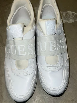 Vita sneakers från Guess - Säljer ett par vita sneakers från Guess med breda elastiska band över foten med stor logotyp. Skorna har präglade mönster i lädret och guldfärgade detaljer på hälen. Perfekt för dig som gillar stilrena och trendiga sneakers.