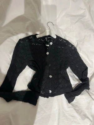 Svart virkad kofta med silvriga knappar - Svart virkad kofta från H&M med snyggt hålmönster och silvriga knappar framtill. Koftan har rund halsringning och långa ärmar, perfekt att slänga över en topp. Materialet är mjukt och lätt, vilket gör den skön att bära.