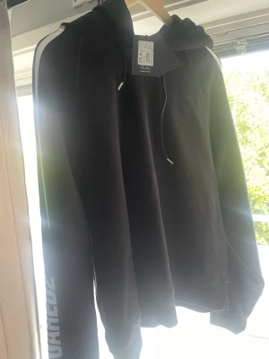 Dsq2 hoodie med svart vita detaljer - Snygg svart hoodie från Dsquared 2, helt ny med prislapp!!!  vita ränder längs axlarna och tryck på ena ärmen. Klassisk huva med dragsko och ribbade muddar vid ärmslut och nederkant. Perfekt för en avslappnad streetwear-look. Helt 