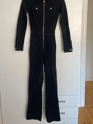 Svart denim jumpsuit med dragkedja - Cool svart jumpsuit i denim med långa ärmar och hel dragkedja framtill. Två bröstfickor med knappar och klassiska jeansdetaljer. Perfekt för dig som gillar en stilren look.