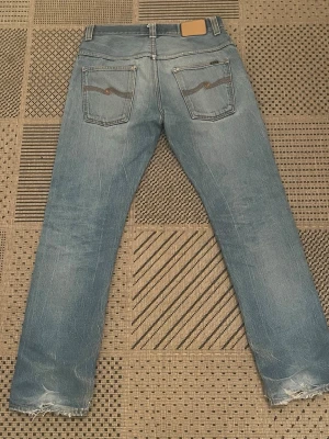 Ljusblå slitna jeans från Nudie - Ljusblå jeans med slitningar och fransade benslut från Nudie Jeans. Klassisk femficksmodell med raka ben och snygga detaljer på bakfickorna. Perfekta för en avslappnad och trendig look. Storlek 30/32