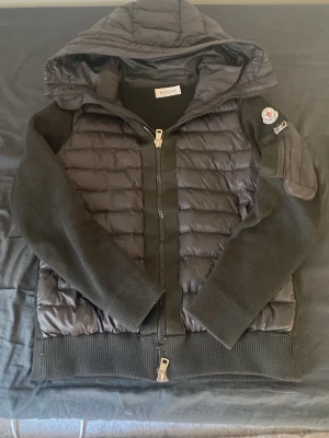 Svart pufferjacka från Moncler - Snygg svart pufferjacka från Moncler med quiltad front och rygg, stickade ärmar och muddar samt huva. Jackan har dragkedja framtill och Moncler-logga på ärmen. Perfekt för kalla dagar och riktigt trendig look.