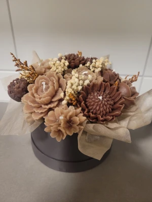 Bukett med blomformade ljus - Unik bukett med handgjorda blomformade ljus i olika nyanser av brunt och beige, arrangerade i en rund mörk ask. Ljusen är detaljerade och ser ut som blommor, blandat med torkade växter för en dekorativ look. Perfekt som inredningsdetalj eller present. Stl m på boxen