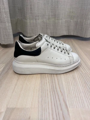 Vita sneakers med svart häl Alexander McQueen - Stilrena vita sneakers från Alexander McQueen med svart detalj på hälen och chunky sula. Skorna har klassisk snörning, perforerade sidor och är tillverkade i läder. Perfekta för dig som gillar en clean och modern look. De är använda men ändå i fint skick, speciellt för priset (sänkte de då sulan är trasig). De är äkta. För ett tag sedan vart de upprustade med vit kulör vid repor. La priset på 980kr men de är köpta för 5000kr