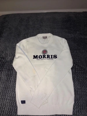Vit stickad tröja från Morris - Vit stickad tröja från Morris med rund halsringning och broderad logga samt brittisk flagga på bröstet. Tröjan har långa ärmar och ribbstickade muddar vid ärmslut och nederkant. Perfekt för en klassisk och stilren look. Nypris 1500-2000kr. En fläck på höger arm som säkert går bort om man har på något medel