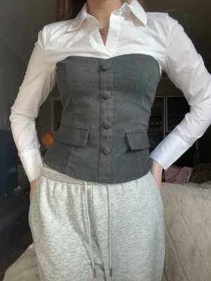 Mörkgrå figursydd skjorta med en låtsasväst - Stilren mörkgrå väst i figursydd modell med knappar framtill och två dekorativa fickor. Västen har en klassisk look och passar perfekt över en skjorta för en dressad stil. Tillverkad i ull för en mjuk och exklusiv känsla.