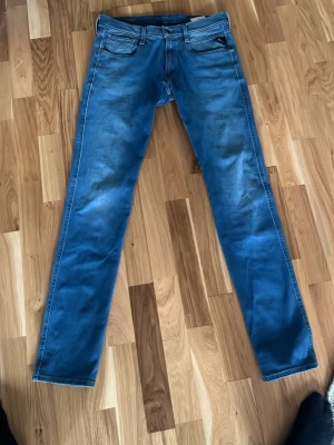Blå Replay Anbass jeans straight fit - Säljer ett par blå Replay Anbass jeans med klassisk femficksdesign och raka ben. Jeansen har snygga slitningar och kontrastsömmar, samt Replay-logga på myntfickan. Perfekta för dig som gillar en tidlös och clean look. Helt oanvända