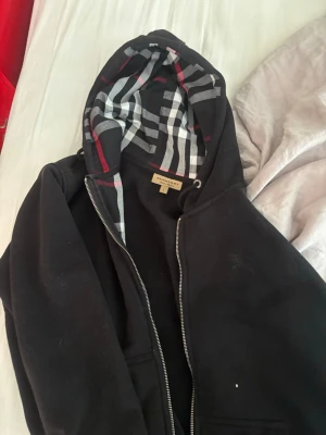 Svart hoodie från Burberry - Säljer en svart hoodie från Burberry med dragkedja och klassiskt rutigt foder i huvan. Hoodien har fickor framtill och justerbar dragsko i huvan. Perfekt för dig som vill ha en stilren och lyxig look med ikoniskt mönster. ÄKTA. Storlek L men passar även bra i M. Pris kan diskuteras😊