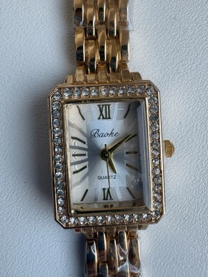 Guldfärgad klocka med stenar - Stilren guldfärgad klocka från Baoke med rektangulär urtavla och gnistrande stenar runt ramen. Klockan har en länkad metallrem och vita detaljer på urtavlan med romerska siffror. Perfekt accessoar för dig som gillar bling och klassisk design.