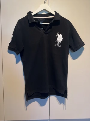 Svart pikétröja från US Polo Assn. - Klassisk svart pikétröja från US Polo Assn med vit broderad logga på bröstet. Tröjan har krage, korta ärmar och knappar vid halsen. Perfekt för en stilren och sportig look.