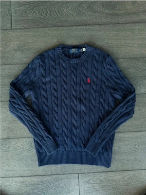 Mörkblå kabelstickad tröja Polo Ralph Lauren - Kabelstickad mörkblå tröja från Polo Ralph Lauren med rund halsringning och ribbade muddar. Tröjan har det klassiska röda Polo-logotypen broderad på bröstet. Perfekt för dig som gillar tidlös och stilren design.