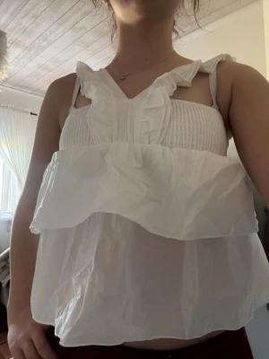Vit volangig linnetopp med smock - Supersöt vit linnetopp med volanger och smockad överdel. Toppen har breda axelband med volangdetaljer och är ärmlös. Perfekt för en fräsch och luftig look under varma dagar.