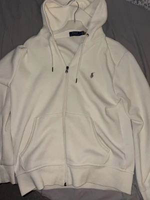 Beige hoodie från Polo Ralph Lauren - Klassisk vit/beige-ish zip-hoodie från Polo Ralph Lauren med dragkedja framtill, två stora fickor och justerbar huva med snören. Liten broderad logga på bröstet. Tillverkad i mjukt bomullsmaterial som känns skönt mot huden. Hoodien är knappast använd och har bara haft på mig den runt 5 gånger. Inga defekter alls och i princip helt ny. Hör av dig vid minsta fundering!!