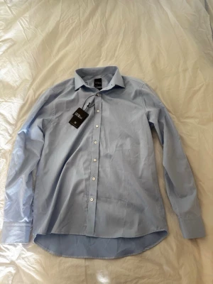 Ljusblå skjorta från Oscar Jacobson - Snygg ljusblå skjorta från Oscar Jacobson i bomull. Klassisk modell med krage och vita knappar framtill. Den är helt ny, dvs aldrig använd. Hör av dig vid frågor!