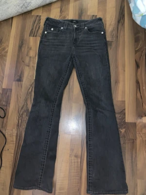 Svarta bootcut jeans True Religion - Säljer ett par svarta bootcut jeans från True Religion med klassiska kontrastsömmar och dekorativa fickor bak med knappar. Jeasen är tillverkade i denim med en lätt tvättad look. Perfekta för en avslappnad men ändå trendig stil.