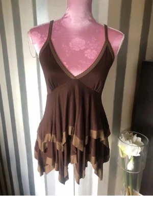 Brun volang linnetopp med smala band - Snygg brun linnetopp med tunna axelband och v-ringning. Toppen har flera lager volanger i olika bruna nyanser och ett flowy, asymmetriskt fall. Perfekt för dig som gillar boho-vibbar och vill ha något unikt till sommaren.