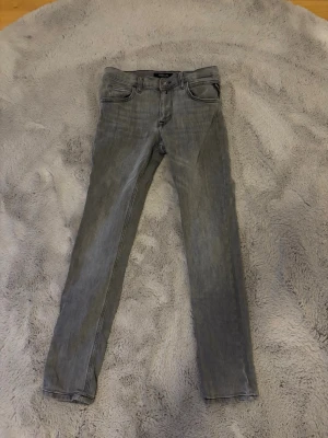 Grå slim fit jeans från Replay - Snygga grå slim fit jeans från Replay med klassisk femficksdesign och diskreta slitningar. Jeansen har en läderpatch med logga bak i midjan och coola detaljer på bakfickorna. Perfekta för en trendig och avslappnad stil.