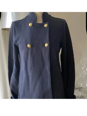 Mörkblå skepparkavaj med guldknappar - Stilren mörkblå skepparkavaj med hög krage och dubbla rader guldknappar framtill. Kavajen har fickor och dekorativa knappar vid ärmsluten. Klassisk rak passform och tillverkad i ett mjukt tyg som ger en snygg siluett.