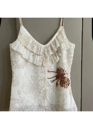 Vit linnetopp med volang och paljetter - Supersöt vit linnetopp med tunna axelband, volang vid ringningen och broderat blommönster. Framsidan har en dekorativ detalj med paljetter i kopparfärg. Perfekt för dig som gillar romantisk och unik stil.