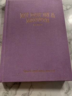 Lila dagbok My Day In A Minute - Snygg lila dagbok med guldfärgad text på omslaget: 'My Day In A Minute (or five)'. Perfekt för snabba dagliga reflektioner och tacksamhetsövningar. Inuti finns färdiga sektioner för dagens lärdomar, tacksamhet och kvällstankar. Hård pärm i tygmaterial.